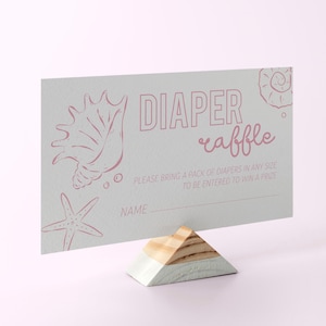 May include: A light blue diaper raffle card with pink seashell and starfish illustrations. The card reads "DIAPER raffle" and includes a line for the name. It is displayed on a wooden stand.