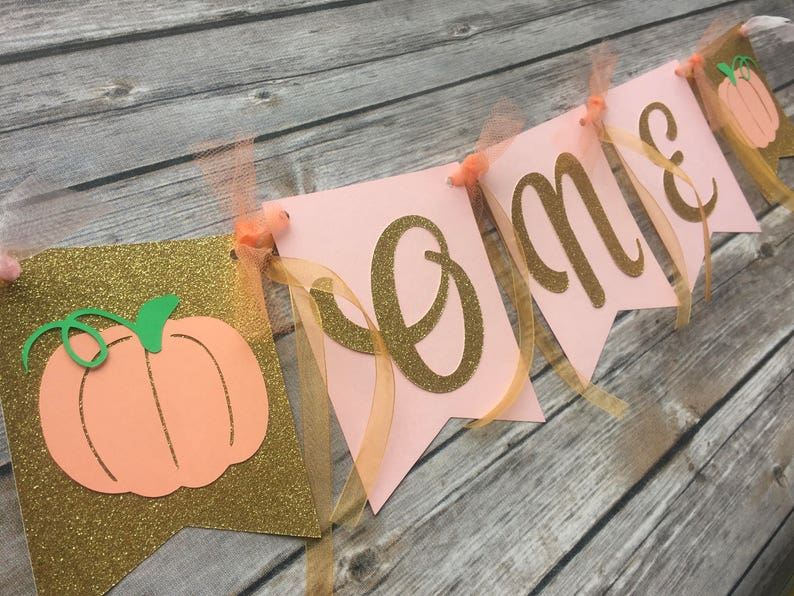 K&ouml;nnte beinhalten: Ein rosa-goldenes Banner mit dem Wort "Home" in goldenen Glitzerbuchstaben. Das Banner ist mit pfirsichfarbenen Filz-K&uuml;rbissen und goldenem Band verziert.