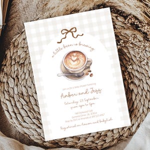 Può includere: Invito per baby shower con un'illustrazione di tazza di caffè con un disegno a forma di cuore nella schiuma. L'invito ha uno sfondo a motivo gingham e il testo "a little bean is brewing". L'invito è per Amber e Jeff.