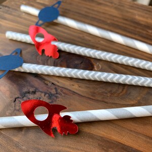 May include: Four gray and white striped paper straws with red rocket and blue planet paper cutouts attached. The straws are on a wooden surface.
