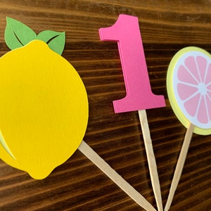 May include: Three cupcake toppers featuring a yellow lemon, a pink number one, and a yellow and pink lemon slice. The toppers are made of paper and have wooden sticks.