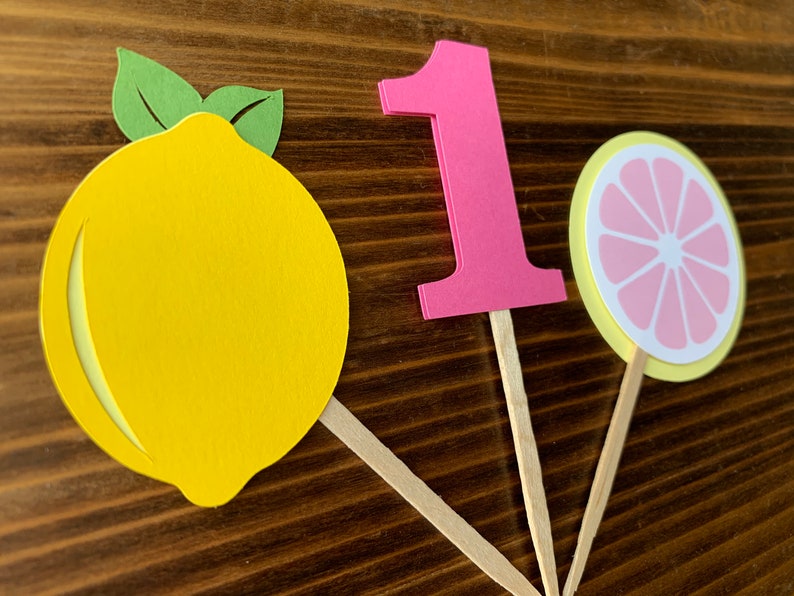 May include: Three cupcake toppers featuring a yellow lemon, a pink number one, and a yellow and pink lemon slice. The toppers are made of paper and have wooden sticks.