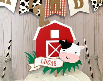 May include: Red barn cake topper with a white cow peeking out, green grass, and a brown banner with the name Lucas. The topper is decorated with a brown number 2.