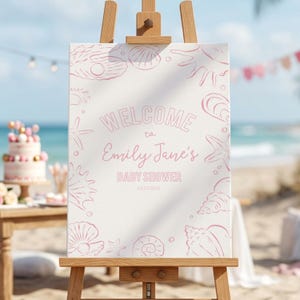 May include: A white welcome sign on a wooden easel, decorated with pink seashell illustrations. The sign reads "WELCOME to Emily Jane's BABY SHOWER 04.23.2026". A cake and decorations are visible in the background.