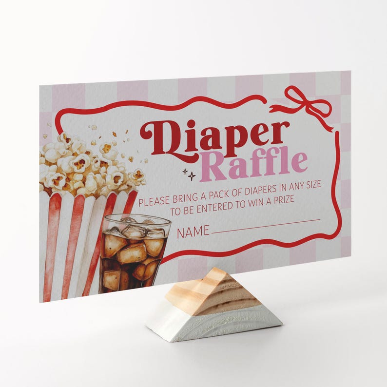 May include: A baby shower raffle card with the words "Diaper Raffle" in a decorative font. The card features a popcorn and soda graphic, with a red and white striped popcorn container and a glass of soda with ice. The card is supported by a wooden stand.