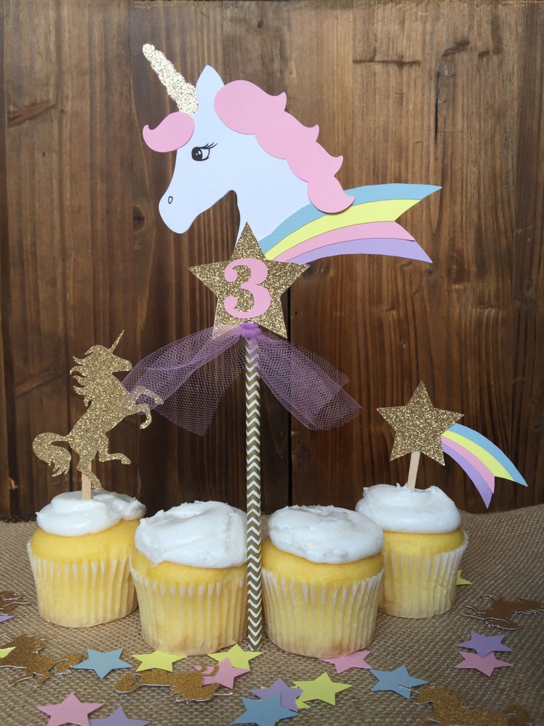 May include: Unicorn cupcake toppers with a rainbow, star, and the number 3. The toppers are on cupcakes with white frosting and are surrounded by confetti.