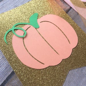 K&ouml;nnte beinhalten: Ein ausgeschnittener Pfirsichfarbener K&uuml;rbis mit einem gr&uuml;nen Stiel auf einem goldenen Glitzerhintergrund. Der K&uuml;rbis ist Teil eines Banners.