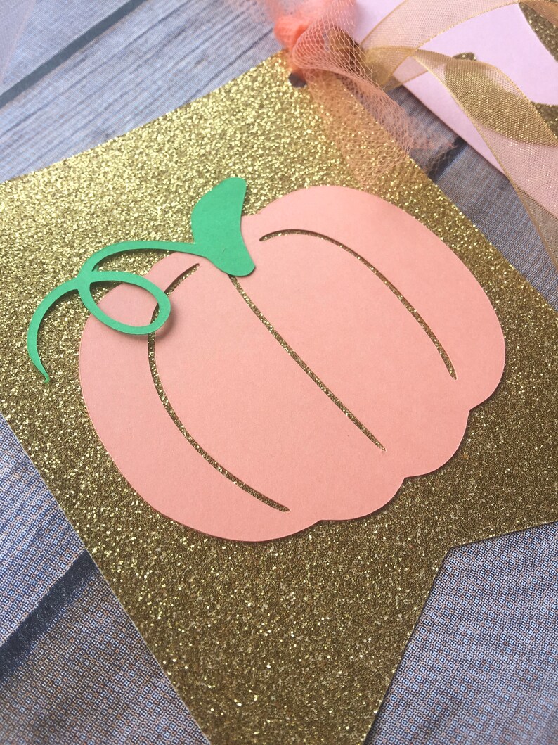 K&ouml;nnte beinhalten: Ein ausgeschnittener Pfirsichfarbener K&uuml;rbis mit einem gr&uuml;nen Stiel auf einem goldenen Glitzerhintergrund. Der K&uuml;rbis ist Teil eines Banners.