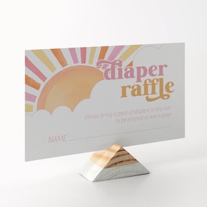 May include: A baby shower diaper raffle card with a retro design. The card features a sun graphic in shades of pink, orange, and yellow, with the words "diaper raffle" in a playful font. The card is held by a wooden stand.