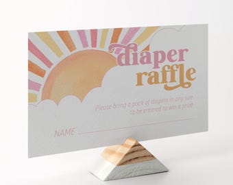 May include: A baby shower diaper raffle card with a retro design. The card features a sun graphic in shades of pink, orange, and yellow, with the words "diaper raffle" in a playful font. The card is held by a wooden stand.