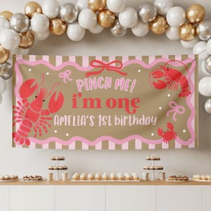 May include: A birthday banner with a tan background, pink border, and red lobster illustrations. The banner reads "Pinch Me! I'm One" and "Amelia's 1st birthday" in pink and red lettering. Balloons and desserts are in the background.