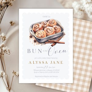 Może przedstawiać: Zaproszenie na baby shower z akwarelową ilustracją bułek cynamonowych w brytfannie. Tekst brzmi "Bun in the Oven" i zawiera imię Alyssa Jane, datę, godzinę i miejsce.