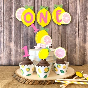 May include: A lemon-themed first birthday cake with pink and yellow decorations. The cake is decorated with a banner that says "ONE" and has a lemon slice on each side. There are also cupcakes with lemon-themed wrappers and a pink number one topper.