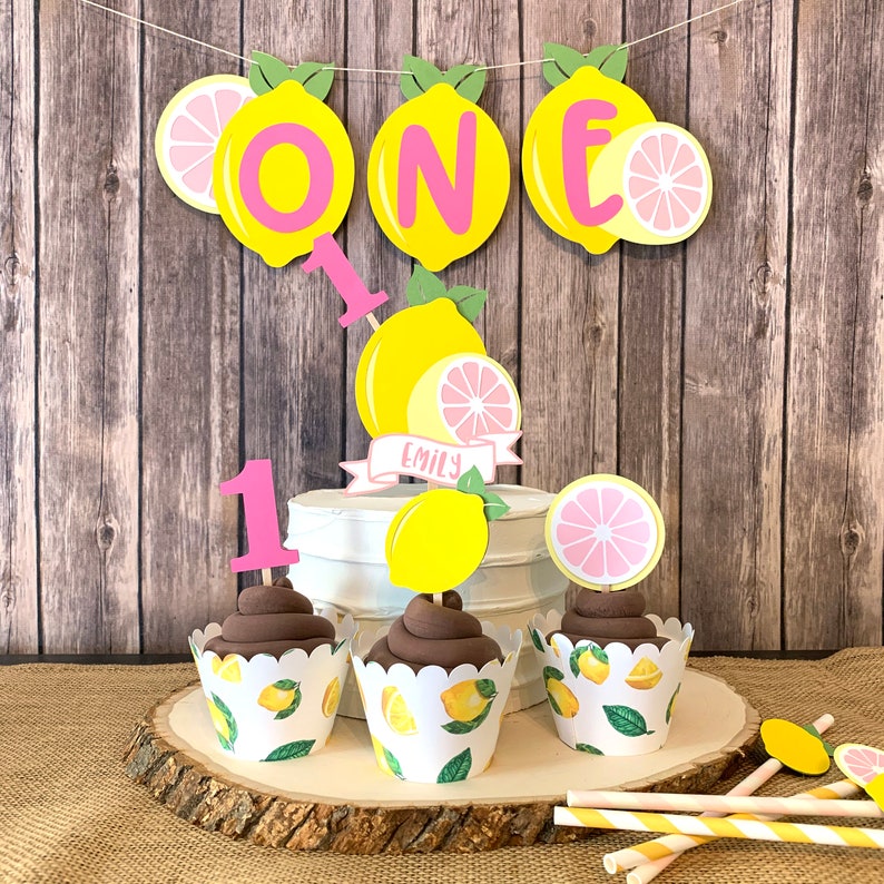 May include: A lemon-themed first birthday cake with pink and yellow decorations. The cake is decorated with a banner that says "ONE" and has a lemon slice on each side. There are also cupcakes with lemon-themed wrappers and a pink number one topper.
