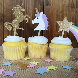 May include: Three cupcakes decorated with unicorn and star themed toppers. The cupcakes are frosted with white frosting and have yellow cake. The toppers are made of glitter paper and feature a gold unicorn, a white unicorn with a purple mane, and a gold star with a rainbow.