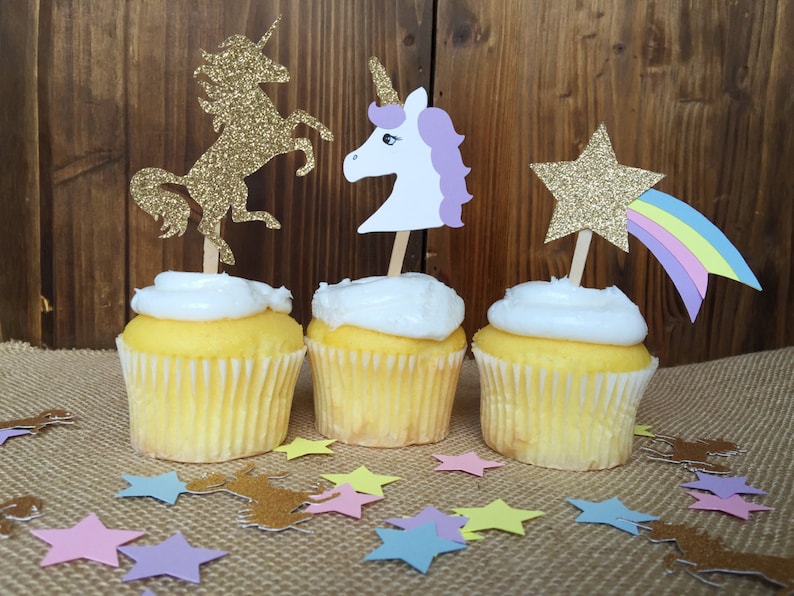 May include: Three cupcakes decorated with unicorn and star themed toppers. The cupcakes are frosted with white frosting and have yellow cake. The toppers are made of glitter paper and feature a gold unicorn, a white unicorn with a purple mane, and a gold star with a rainbow.