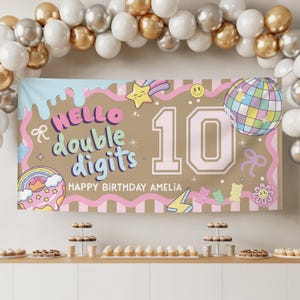 May include: A birthday banner with the text "HELLO double digits" and "HAPPY BIRTHDAY AMELIA". The banner features a number 10, a disco ball, a shooting star, a rainbow donut, and other colorful graphics. Balloons in gold, silver, and white are in the background.