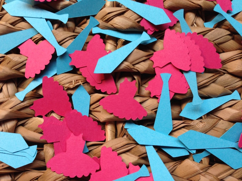 Pode incluir: Uma cole&ccedil;&atilde;o de recortes de papel azul e rosa em forma de gravatas e vestidos. Os recortes est&atilde;o espalhados sobre uma superf&iacute;cie tecida.