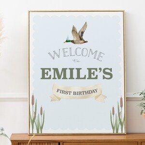 Könnte beinhalten: Gerahmter Druck mit hellblauem Hintergrund und gewelltem Rand. Der Druck zeigt eine fliegende Ente mit Partyhut über den Worten "WELCOME to EMILE'S FIRST BIRTHDAY". Unten sind Rohrkolben und Schilf abgebildet.