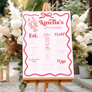 May include: A pink and white birthday sign on an easel. The sign says "Amelia's first birthday" and includes details like weight (21 lbs), height (29 inches), and favorite things. A lobster illustration is at the top.