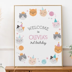May include: A white welcome sign with a pink border and a circle of cute cartoon cats wearing party hats. The sign says "Welcome to Olivia's 3rd birthday".
