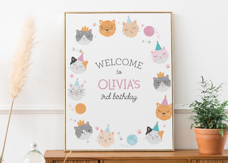 May include: A white welcome sign with a pink border and a circle of cute cartoon cats wearing party hats. The sign says "Welcome to Olivia's 3rd birthday".
