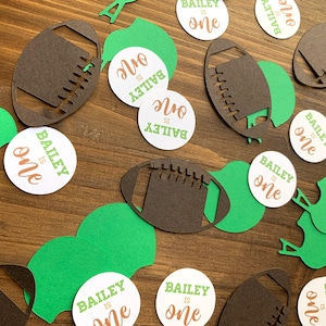 Peut inclure: Des découpes en papier vert et marron de ballons de football et de casques avec des cercles blancs qui disent "Bailey is one" en texte marron.