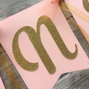 K&ouml;nnte beinhalten: Rosa Banner mit einem goldenen Glitzerbuchstaben "M" darauf. Das Banner hat ein goldenes Band und ein pfirsichfarbenes T&uuml;llband daran befestigt.
