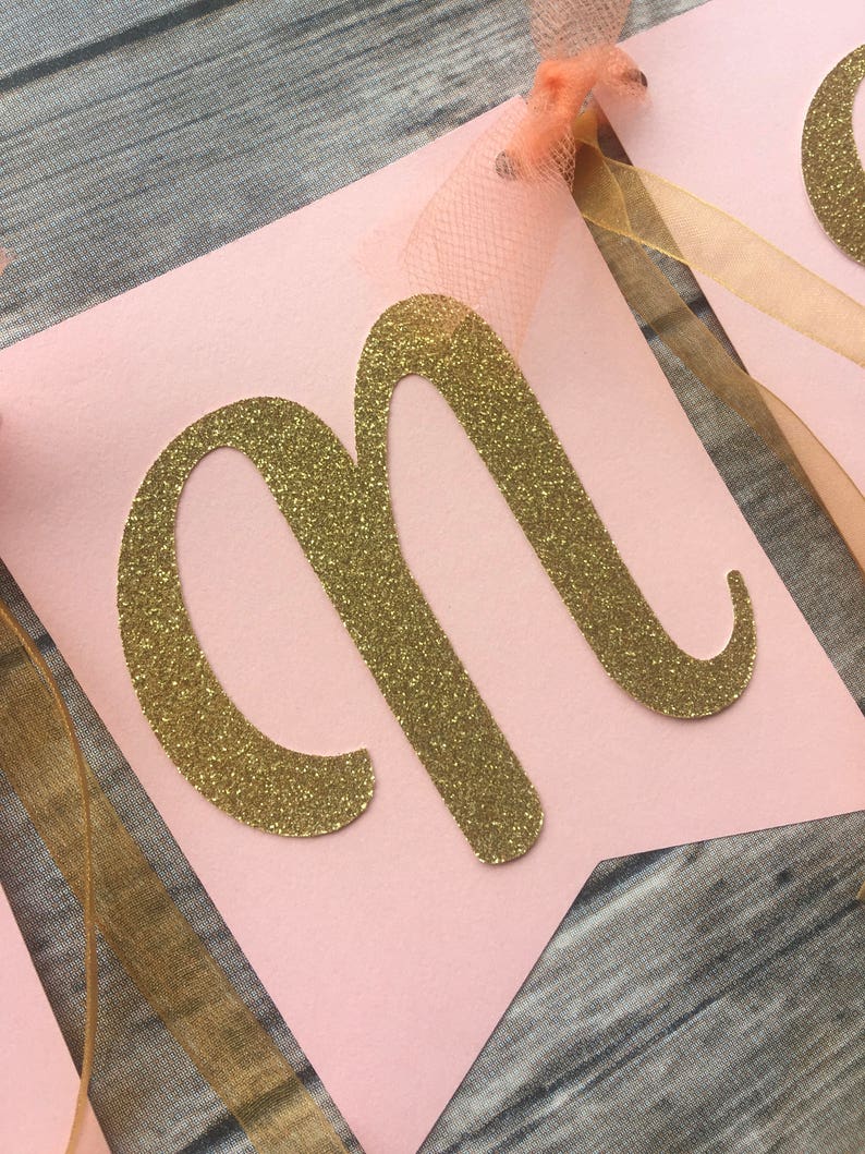 K&ouml;nnte beinhalten: Rosa Banner mit einem goldenen Glitzerbuchstaben "M" darauf. Das Banner hat ein goldenes Band und ein pfirsichfarbenes T&uuml;llband daran befestigt.