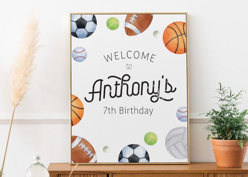 May include: A white sign with a gold frame featuring a watercolor illustration of various sports balls, including a basketball, baseball, football, soccer ball, and tennis ball. The sign reads "Welcome to Anthony's 7th Birthday".