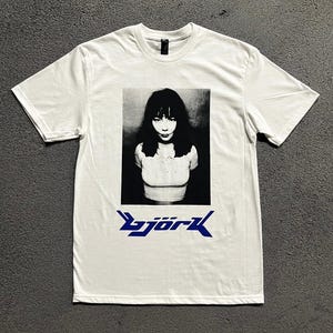 Pode incluir: Uma t-shirt branca de manga curta com uma impressão fotográfica a preto e branco de uma pessoa com cabelo escuro no peito. Abaixo da fotografia, o texto "björk" está impresso numa fonte azul estilizada com letras angulares.