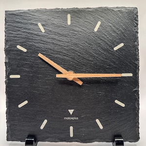 Könnte beinhalten: Eine quadratische Wanduhr aus schwarzem Schiefer mit rauer, strukturierter Kante. Sie verfügt über helle Stundenmarkierungen, dünne Uhrzeiger aus Holz und den in Weiß gedruckten Schriftzug matoxplus unter der Mitte. Die Uhr steht auf zwei schwarzen Ständern.