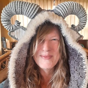 May include: A woman wearing a gray and white crocheted hooded hat with large horns. The hat has a fluffy white trim around the hood.