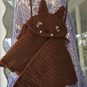 May include: A brown crocheted bat-shaped bag with a strap. The bag has a cute face with white teeth and large eyes.