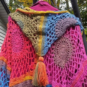 May include: A colorful crocheted shawl with a fringe tassel. The shawl is made of different colored squares with a circular pattern in the center of each square. The colors are pink, blue, green, orange, and brown.