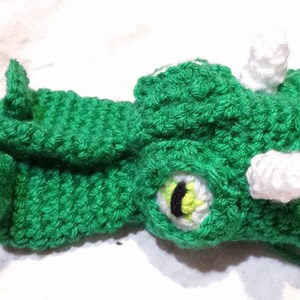 May include: A green crocheted dragon hat with white accents. The hat features a dragon head with a detailed eye, horns, and a loop for securing. The handmade hat is suitable for costume or play.
