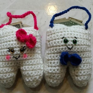 May include: Two crocheted tooth-shaped coin purses with embroidered faces. One purse has a pink bow and the other has a blue bow. Both purses have a loop at the top for hanging.