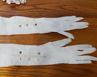 May include: A pair of long, white gloves. The gloves are made of a smooth fabric and extend well past the elbow. Each glove has a decorative band with small, silver-colored studs near the wrist.