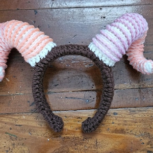 May include: A handmade crochet headband with two curved horns. The horns are pink and white striped, with a white base and tips. The headband itself is brown. This whimsical accessory is perfect for costumes or themed events.