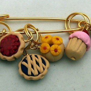 May include: A gold safety pin with six charms attached. The charms are shaped like different pastries, including a chocolate pie, a cherry pie, a lemon pie, a cupcake, and a white cake with a pink rose.