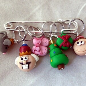 May include: A set of six colorful Christmas-themed stitch markers, including a mouse, a king, a pair of pink shoes, a Christmas tree, a present, and a girl with a red bow. The markers are attached to a silver safety pin.