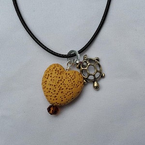 Könnte beinhalten: Eine schwarze Schnurkette mit einem Lava-Stein-Herz-Anhänger, einem kleinen goldenen Schildkröten-Anhänger und einer braunen Kristallperle.