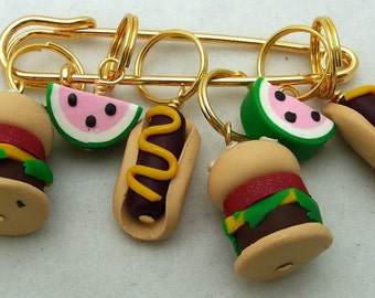 May include: A gold safety pin with five charms: a hamburger, a hot dog, a slice of watermelon, a taco, and another hamburger. The charms are made of polymer clay and are brightly colored.