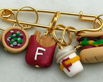 May include: A gold-colored safety pin with miniature food charms. The charms include a hamburger, pizza, french fries, a drink, a sandwich, and a hot dog. The food charms are made of clay and have detailed features.