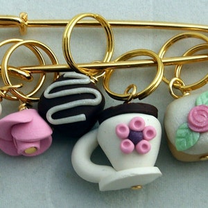 May include: A gold safety pin with six charms attached. The charms are made of white and pink polymer clay and are shaped like a teapot, a rose, a cupcake, a chocolate-covered swirl, and two teacups.