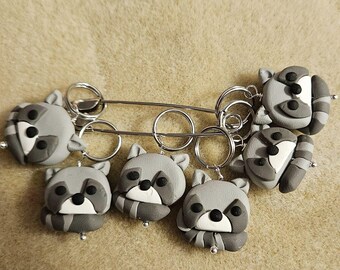 May include: Six gray raccoon stitch markers with black eyes and white noses, attached to a silver metal ring.