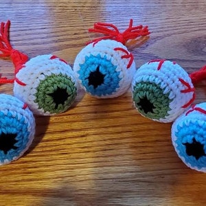 May include: Five crocheted eyeball ornaments with red yarn accents. Each eyeball is white with a blue or green iris and a black pupil.