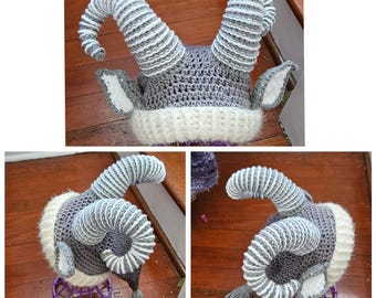 May include: A gray and white crocheted hat with horns and ears. The hat is made with a soft yarn and has a fluffy texture. The horns are curled and the ears are floppy.