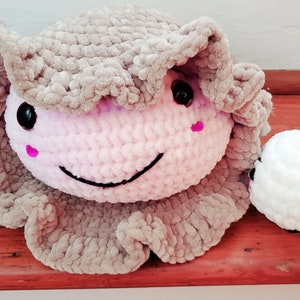 以下が含まれることがあります： 2つの編みぐるみ。大きい方は薄いピンクとグレーで笑顔をしています。小さい方は白で黒い刺繍の顔をしています。