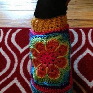 May include: A black dog wearing a colorful, handmade crochet dog sweater. The sweater features a large flower design in the center, with a rainbow of colors including red, orange, green, blue, and yellow. The dog is sitting on a red and white patterned rug.